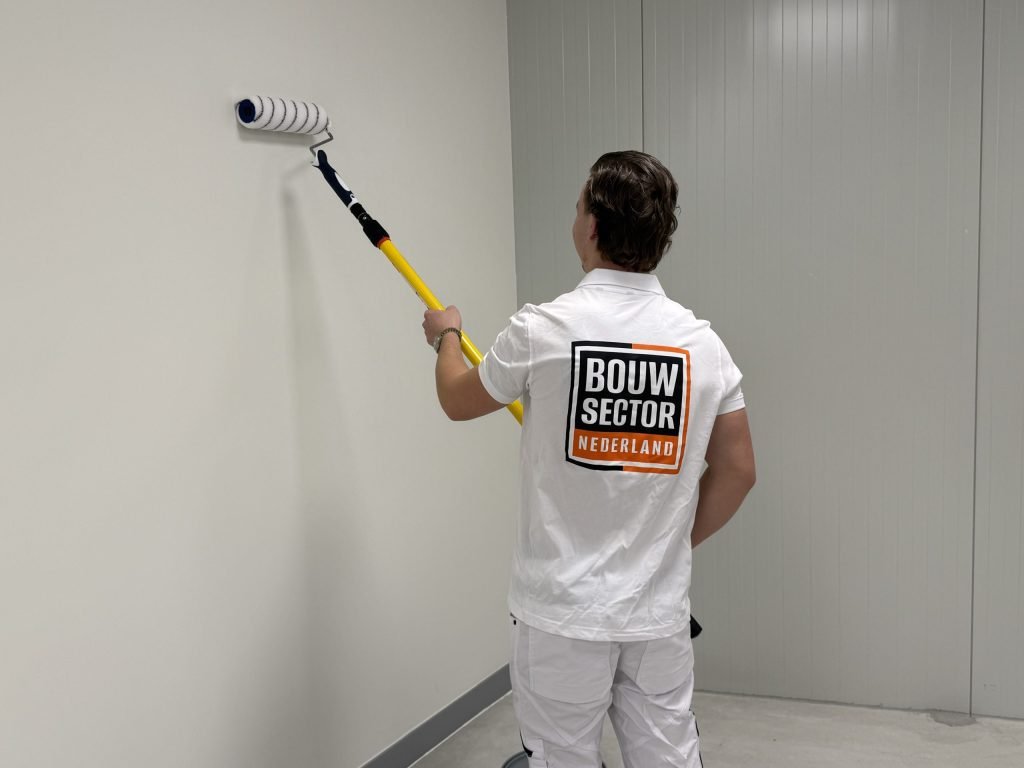 Vakman in witte werkkleding saust een witte wand met een roller aan een verlengsteel