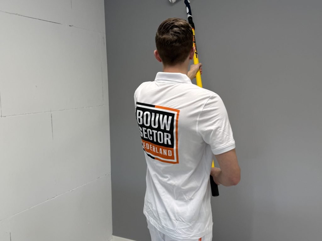Schilder werkt een grijze wand af met een roller aan een lange steel, naast een witte muur