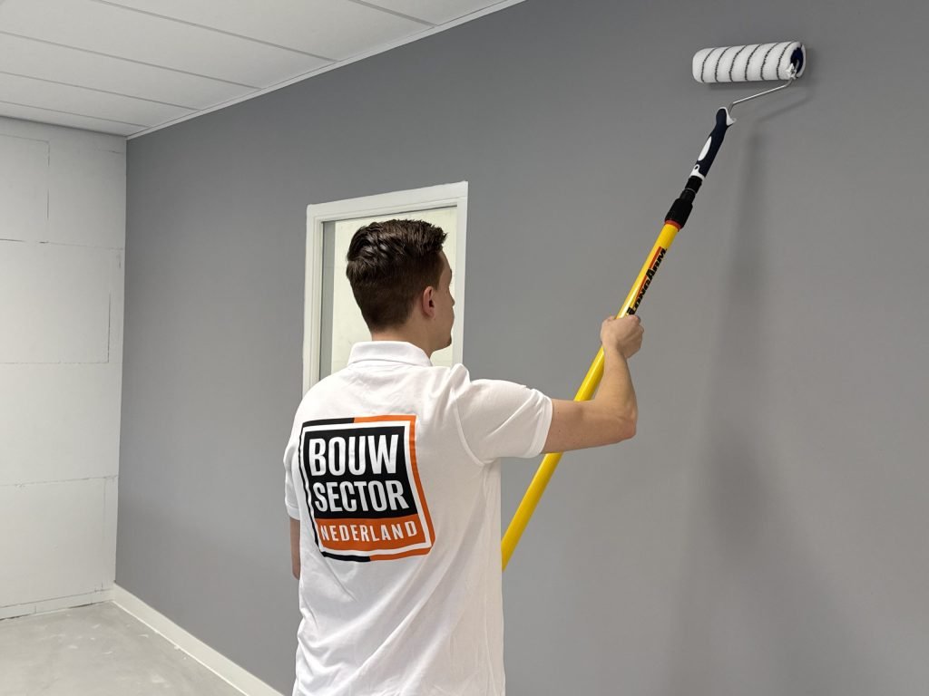 Schilder met het logo van Bouwsector Nederland op de rug rolt een grijze muur naast een wit kozijn