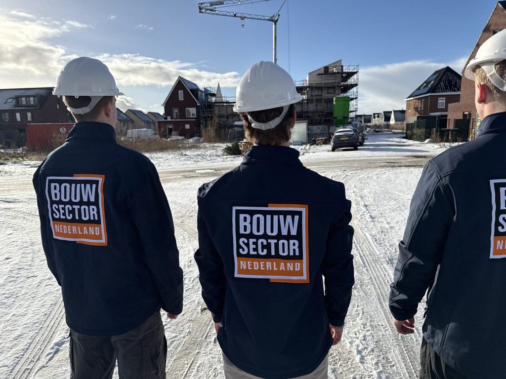 Drie vakmensen van Bouwsector Nederland met witte helm kijken uit over een nieuwbouwwijk in de sneeuw